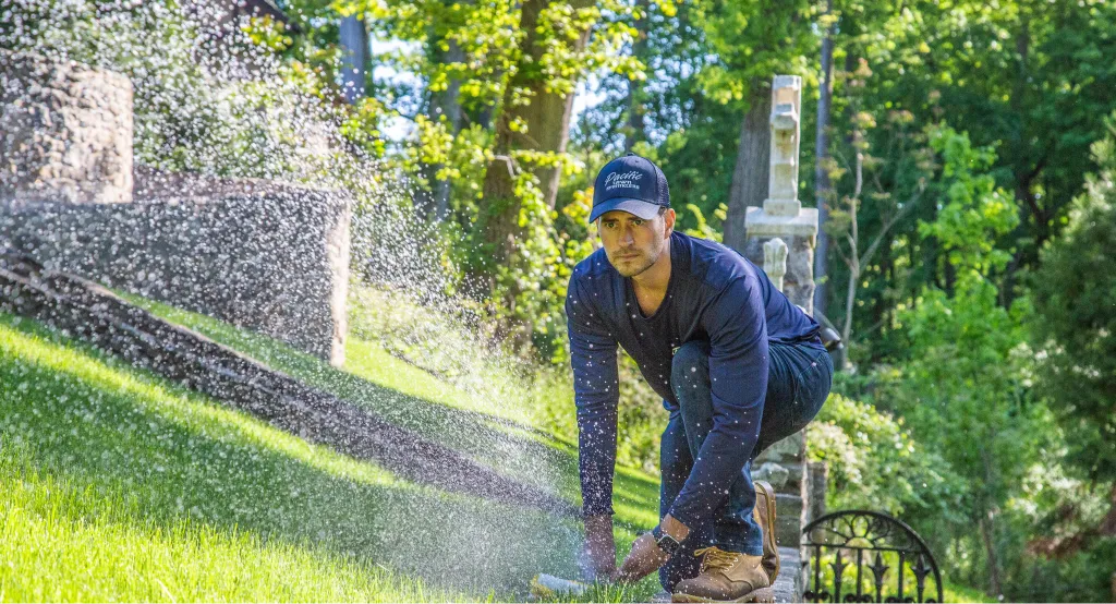 A man wearing a blue cap and shirt crouches by a sprinkler watering a lush green